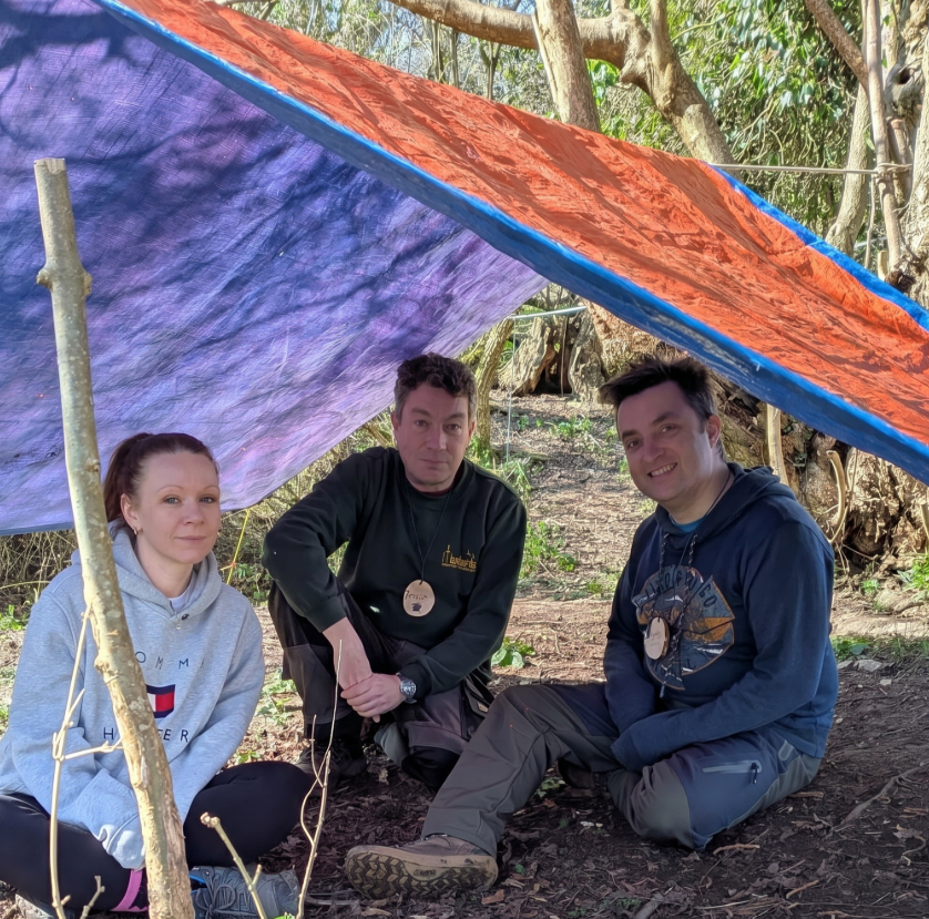 Trainees sitting together under a tarpaulin shelter at the woodland training site